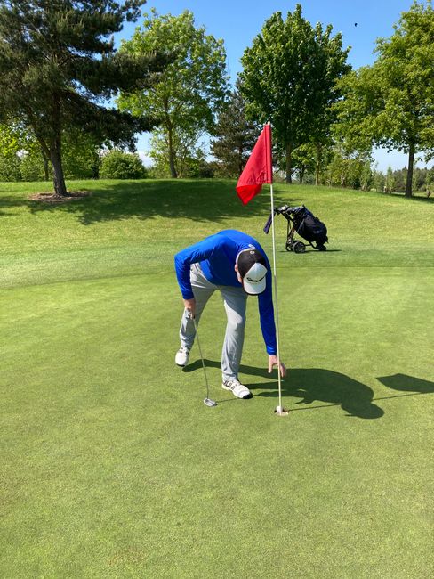 A person wearing a white cap, blue jacket, and light-colored trousers is on a well-maintained golf green at Arkley Golf Club, preparing to putt. They are bending forward with a golf club in hand, near