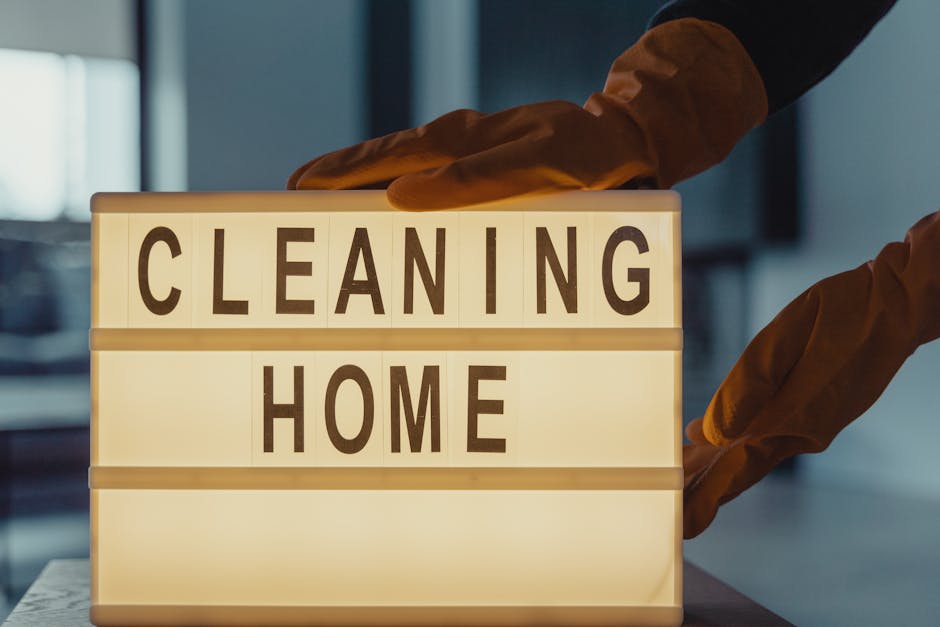 A person wearing orange cleaning gloves is arranging a white illuminated signboard on a wooden surface. The signboard displays the words 'CLEANING HOME' in bold, black letters. The background includes