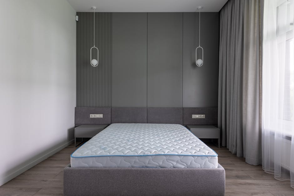 A neatly made bed with a dark fabric headboard in a bedroom featuring a layered arrangement of white and grey pillows and bedding. The bed is covered with a set of grey and white bed linens, appearing